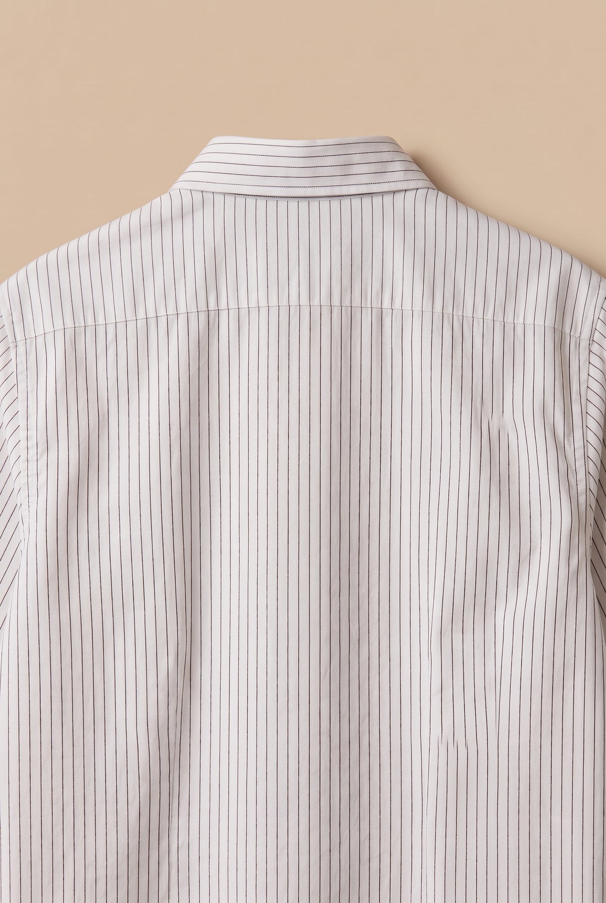 Close-up of the back of a white long-sleeve button-down shirt with thin vertical gray pinstripes, set against a solid beige background.